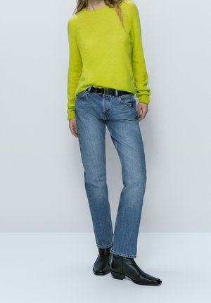Personne portant un pull vert vif, un jean bleu avec une ceinture noire et des bottines noires, debout devant un fond clair uni.