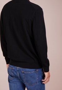 Hombre vestido con un suéter negro de manga larga y jeans azules, de pie de espaldas a la cámara, contra un fondo liso.