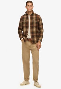 Brunternet jakkesæt med fire knapper og to brystlommer, parret med en lys langærmet skjorte og beige bukser, afsluttet med beige sneakers.