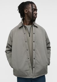 Γκρι, επενδυμένο μπουφάν με κουμπωτό εμπρός, μεγάλο γιακά και πλαϊνές τσέπες, φορεμένο πάνω από γκρι τοπ με φερμουάρ. Απλός, λειτουργικός σχεδιασμός.