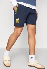 Σορτς ναυτικού με έμβλημα AFA σε κίτρινο, με πλαϊνές τσέπες και αντίθετη πράσινη ρίγα. Φοριέται με λευκές κάλτσες και μπεζ παπούτσια.