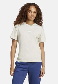 Camiseta de algodón color beige claro con mangas cortas, cuello redondo y pequeño logo blanco en el pecho. Lleva puesto un pantalón de chándal azul.