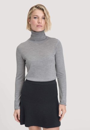 Donna con capelli biondi corti che indossa un maglione a collo alto grigio chiaro e una gonna in maglia nera a coste, in piedi di fronte a uno sfondo chiaro.