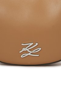 Tan lærveske med glatt tekstur, en sølvfarget logo og sorte detaljer. Minimalistisk design med avrundet form.