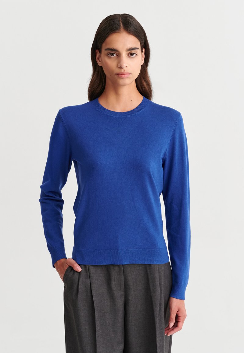 Femme aux longs cheveux foncés portant un pull bleu vif à manches longues et un pantalon ample gris foncé sur un fond uni.
