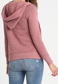 Mujer vistiendo una sudadera rosa polvoriento y jeans skinny rotos de color azul claro, vista desde atrás sobre un fondo blanco liso.