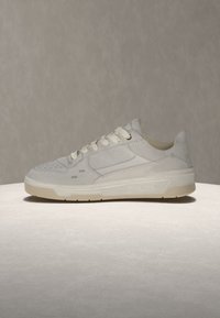 Baskets basses blanches avec lacets sur le devant, panneaux latéraux perforés et semelle beige clair, présentées sur une surface lisse et claire.