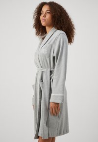 Roupa de banho cinza em tecido suave, com um design de envoltório, cinto e dois bolsos. Iniciais bordadas no bolso do peito acrescentam detalhe.