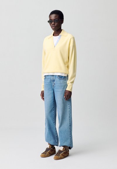 Pulóver de punto amarillo con cuello y detalle de botones, superpuesto sobre una camisa azul claro. Jeans azul claro desgastados y zapatillas marrones.