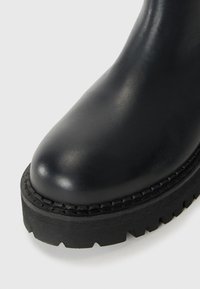 Gros plan sur une botte en cuir noir avec un bout arrondi et une semelle épaisse en caoutchouc robuste sur fond blanc.