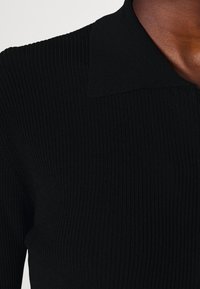 Närbild av en person som bär en svart ribbad stickad topp med krage, som visar textilens struktur och axeldetaljer.