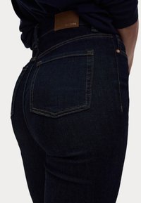 Jean en denim bleu foncé à la coupe ajustée, avec une étiquette en cuir portant "EST. 1884" et des coutures contrastées. Poches arrière visibles.