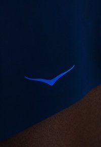 Tummansininen kangas, jossa pieni kaareva sininen logo, joka muistuttaa tyyliteltyä lintua tai valintamerkkiä kulman lähellä ruskealla taustalla.