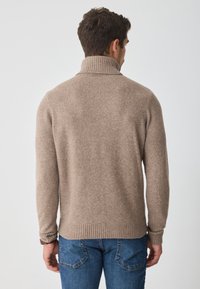 Bezs színű kötött pulóver magas bordás gallérral, hosszú ujjak, és bordás alj, kék farmernadrágon viselve. Puha tapintású, hétköznapi stílusú.