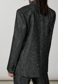 Persona che indossa un blazer nero glitterato e testurizzato con pantaloni abbinati, mostrata di spalle su uno sfondo chiaro e semplice.