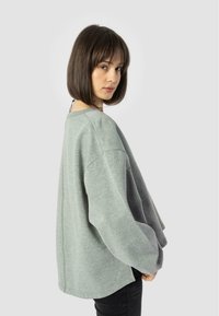 Felpa grigia chiaro con vestibilità ampia, spalle scese e texture morbida. Presenta un collo rotondo e fessure laterali discreti.