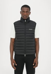 Sort puffer vest med horisontalt quiltet design, høj krave, frontlynlås og et lille logo på venstre side. Sammen med en hvid skjorte.