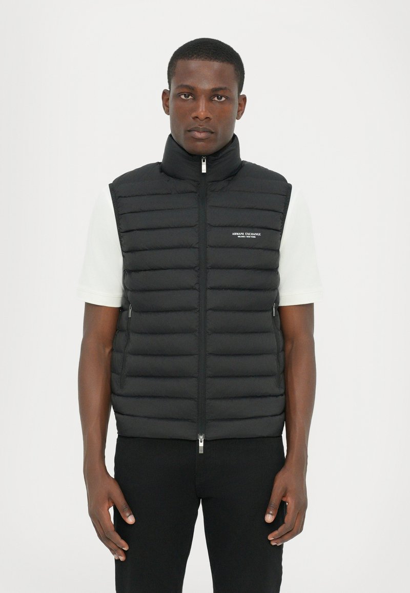 Sort puffer vest med horisontalt quiltet design, høj krave, frontlynlås og et lille logo på venstre side. Sammen med en hvid skjorte.