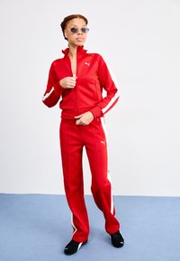 Crveni trenirka s bijelim bočnim prugama, jakna na zip i hlače s elastičnom trakom, uparen s crnim sportskim cipelama i sunčanim naočalama.