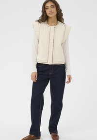 Prošívaná béžová vesta s volánkovými rameny, zapínáním na pět knoflíků, dekorativním prošíváním, kombinovaná s tmavými džínovými kalhotami a hnědými mokasíny.