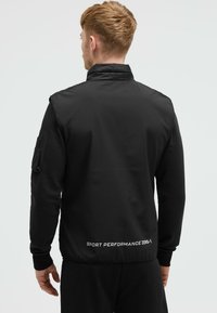 Giacca atletica nera con colletto alto, chiusura con zip, tessuto texturizzato e testo del logo sull'orlo inferiore. Maniche corte con tasca laterale.
