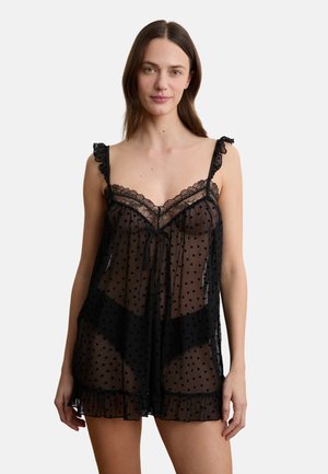 Frau trägt schwarze, transparente Unterwäsche mit Herzmustern, Spitzenbesatz und Rüschenträgern, steht vor einem einfarbigen weißen Hintergrund.