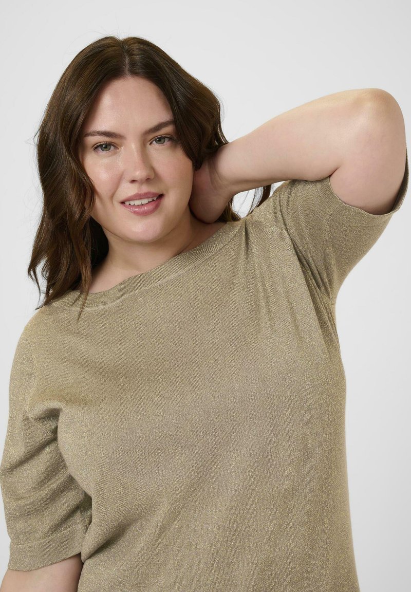 Vrouw met golvend bruin haar, draagt een lichtbruin shirt met korte mouwen, glimlachend met één hand achter haar hoofd tegen een effen achtergrond.