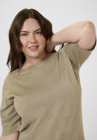 Mujer con cabello castaño ondulado que lleva una blusa de manga corta color marrón claro, sonriendo con una mano detrás de la cabeza contra un fondo liso.