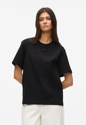 Młoda kobieta o długich brązowych włosach, ubrana w czarną oversize'ową koszulkę i białe spodnie, stoi z rękami za plecami.