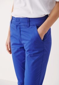 Klart blå byxor i ett mjukt bomullsmaterial, med sidofickor och en skräddarsydd passform, där bärarens hand vilar i en ficka.