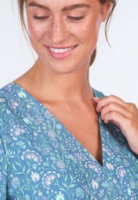 Blumenkleid mit V-Ausschnitt, das einen blauen Hintergrund mit rosa und grünen botanischen Mustern zeigt, aus leichtem Stoff mit weicher Textur gefertigt.