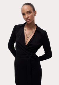 Vestido negro envolvente con mangas largas, escote en v y detalle de cintura anudada. Presenta un collar de cadena dorada y una textura de tela lisa.