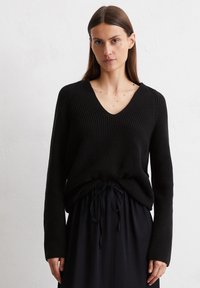 Schwarzer gerippter Pullover mit tiefem V-Ausschnitt und langen Ärmeln, kombiniert mit einem schwarzen Rock mit Kordelzug. Strukturierter Stoff durchgehend.