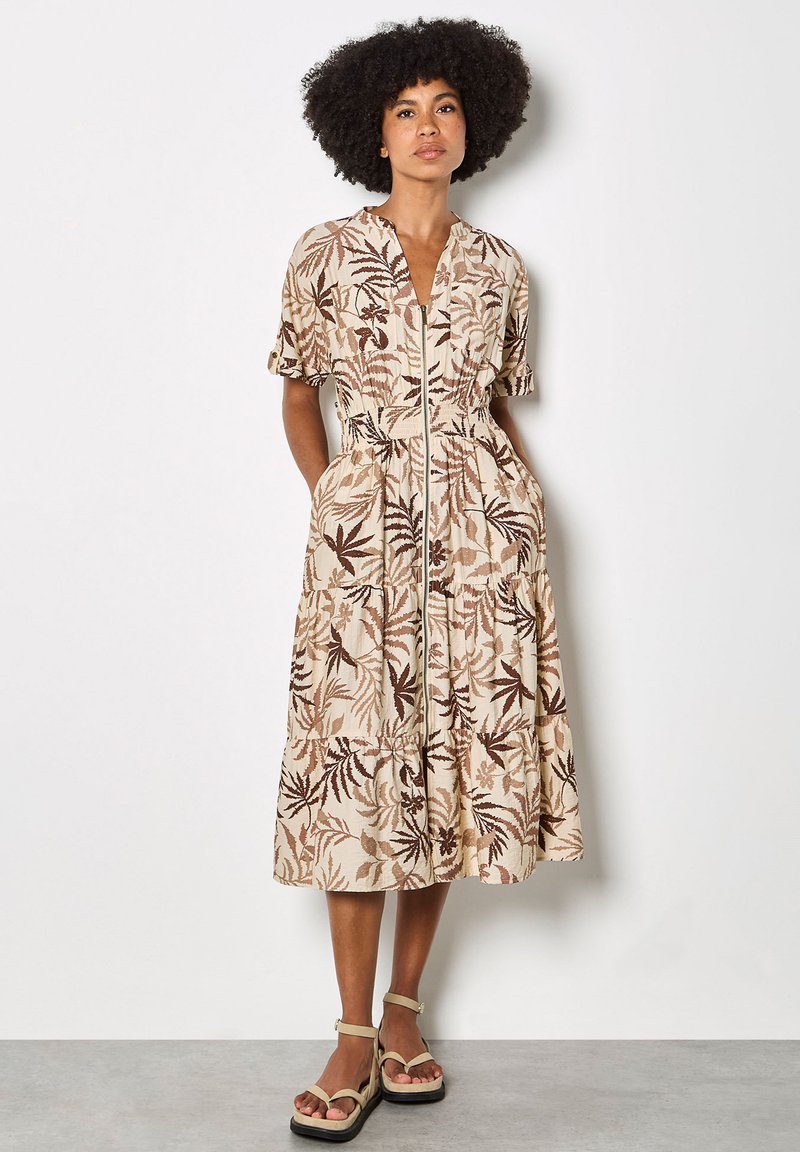 Femme debout portant une robe midi beige avec des motifs de feuilles marron, manches courtes, fermeture éclair à l'avant, et des sandales compensées beige.