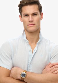 Jasnoniebieska lniana koszula z kołnierzykiem stojącym, krótkimi rękawami i małym logo, w połączeniu ze złotym i granatowym zegarkiem chronografem na nadgarstku.