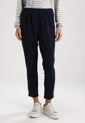 Femme portant un pantalon bleu marine ajusté, un haut à manches longues rayé gris et blanc, et des baskets blanches, debout devant un fond uni.