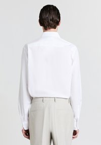 Chemise blanche à manches longues avec un col classique, associée à un pantalon gris clair. Tissu lisse, coupe sur mesure et poignets boutonnés.