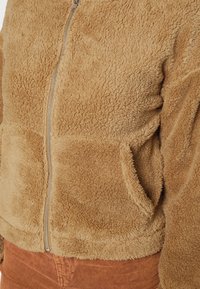 Giacca in fleece marrone con chiusura a zip, texture morbida e tasche frontali. Il design è corto con un orlo arrotondato.