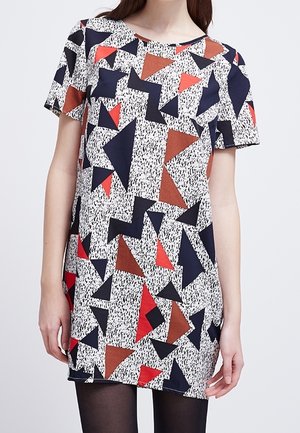 Femme portant une robe à manches courtes avec des motifs géométriques de triangles noirs, rouges et bruns sur un fond blanc tacheté.