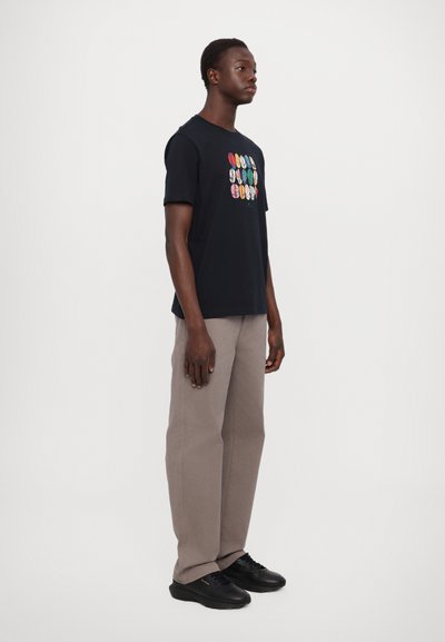 T-shirt noire à manches courtes avec des graphiques circulaires colorés sur le devant, associée à un pantalon marron clair et des baskets noires.