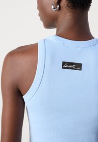 Personne à la peau foncée portant une boucle d'oreille argentée en forme d'anneau et un débardeur bleu clair côtelé avec une petite étiquette noire rectangulaire au dos.