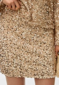 Krótką dopasowaną sukienkę w jasnym beżu pokrytą cekinami o różnych rozmiarach, z gładką teksturą i lekko drapowaną sylwetką.
