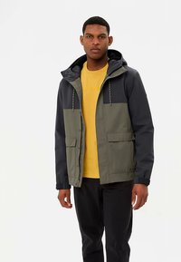 Herrenjacke in Dunkelblau und Olivgrün. Verfügt über eine Kapuze, Fronttaschen und eine Knopfleiste. Wird über einem gelben Hemd getragen.