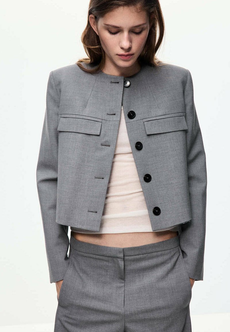 Jeune femme regardant vers le bas porte une veste de costume courte gris clair avec des boutons et un pantalon assorti, avec un top beige clair en dessous.