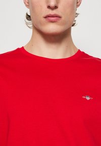 T-shirt rouge à manches courtes en tissu lisse avec col rond. Présente un petit logo blanc sur la poitrine. Vue en gros plan.