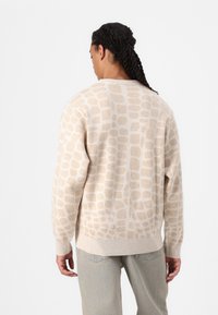 Beiger Pullover mit einem strukturierten, hellbraunen, abstrakten Muster. Gerippte Säume und ein lockerer Schnitt, kombiniert mit hellblauen Jeans.