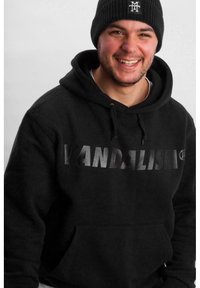 Black pullover hoodie featuring "VANDALISM" in glossy print, soft fabric, and a front pocket. Styled with a black ribbed beanie.