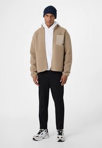 Hellbraune Fleecejacke mit einer beigefarbenen Brusttasche, weißem Hoodie darunter, schwarzen Hosen und marineblauer Strickmütze. Weiße und schwarze Sneakers vervollständigen das Outfit.