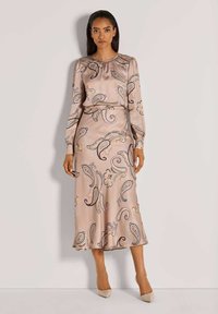 Langes mauvefarbenes Satin-Kleid mit einem Paisleymuster in Blau und Schwarz, langen Ärmeln und einem fließenden Rock. Kombiniert mit nudefarbenen High Heels.