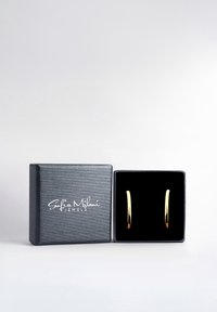 Orecchini a cerchio dorati adagiati in una scatola foderata in velluto nero con un esterno grigio texturizzato e logo bianco. Design semplice ed elegante, finitura liscia.